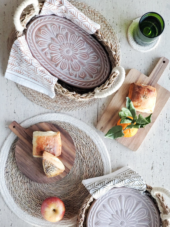 Terracotta plates engraved with a flower design into an oval and a round basket, hand screen-printed tea towels printed in two colors on natural cotton, Wooden serving boards, Savar placemat, Kata placemat and coasters